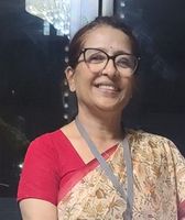 Smiling woman in glasses and a floral sari stands indoors, wearing a red blouse and a lanyard.
