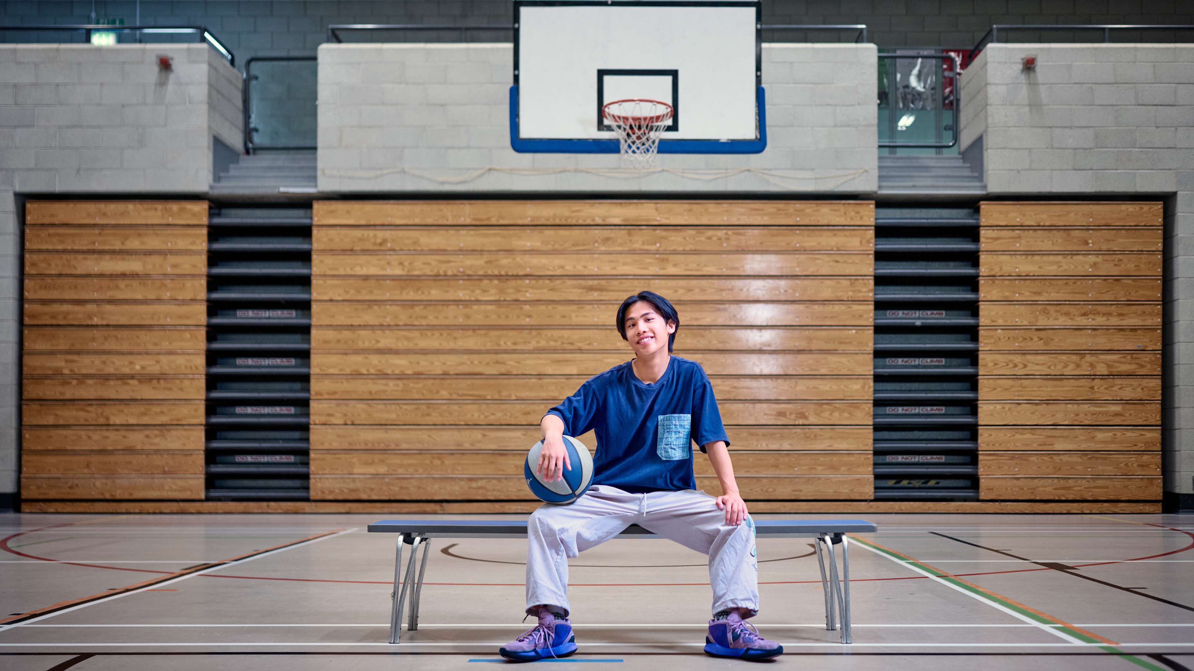 A student sits on a bench holding a basketball. He is in a gym and a basketball net is behind him.