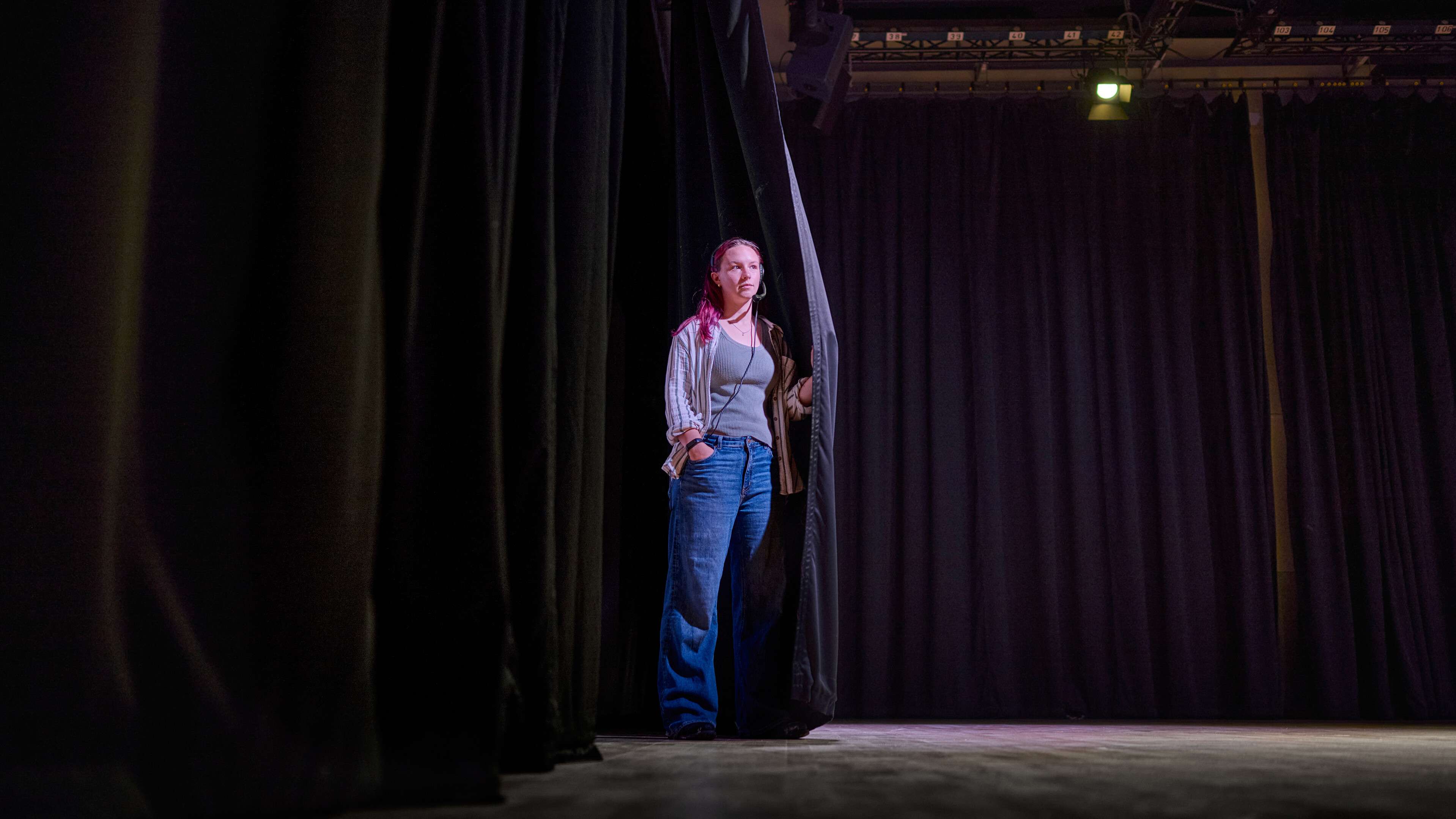 A student stands in the wings of a stage. They have a headset on.
