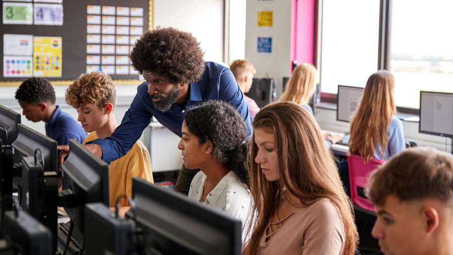 Male Teacher Helping Teenage Female High School Student Working In Computer Class