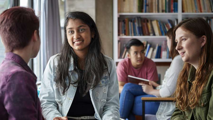 Two groups of students chatting in a seminar room. They are looking at textbooks and talking, and there is a wall full of bookshelves behind them.