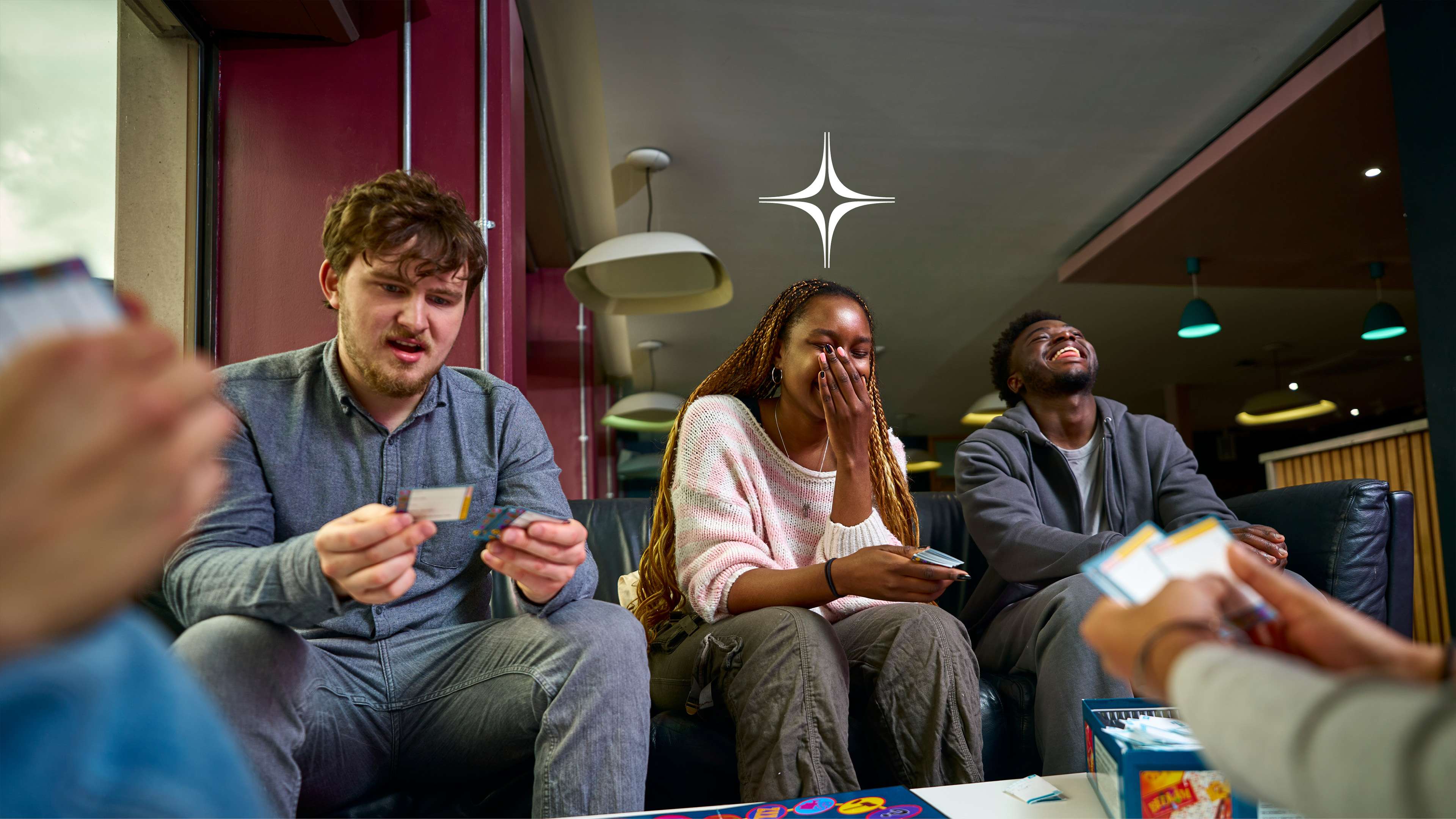 A group of university students laughing and enjoying a card game together in a modern student common room.