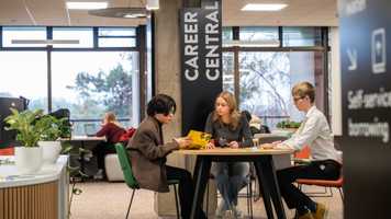 Three students sitting at a table at the UEA careers centre, discussing career opportunities while holding yellow brochures. A sign reading 'Career Central' is visible in the background, along with other students working on laptops in a modern, well-lit space.