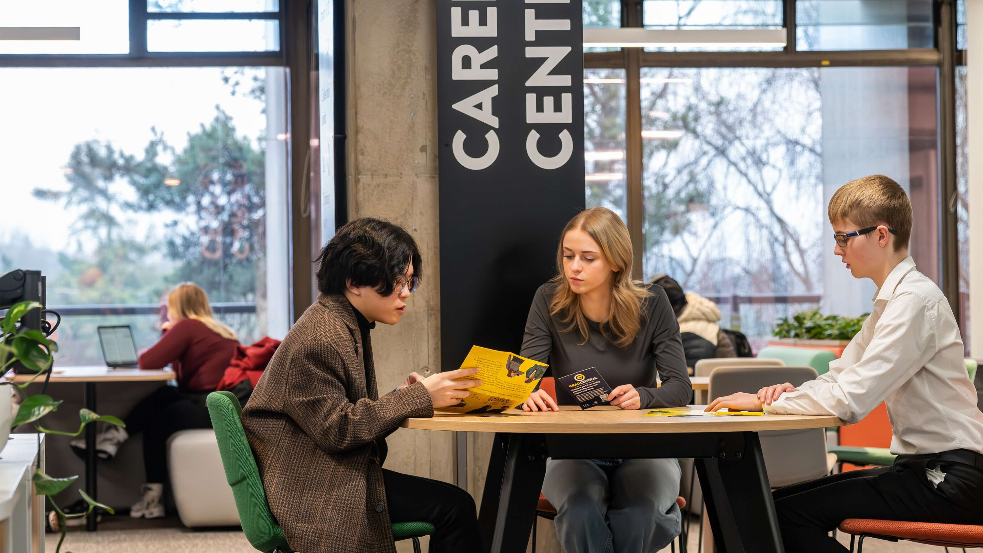 Three students sitting at a table at the UEA careers centre, discussing career opportunities while holding yellow brochures. A sign reading 'Career Central' is visible in the background, along with other students working on laptops in a modern, well-lit space.