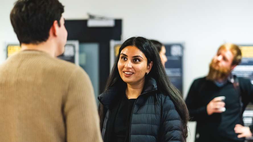 Three people engaged in conversation indoors; a woman in the center smiles, wearing a black jacket.