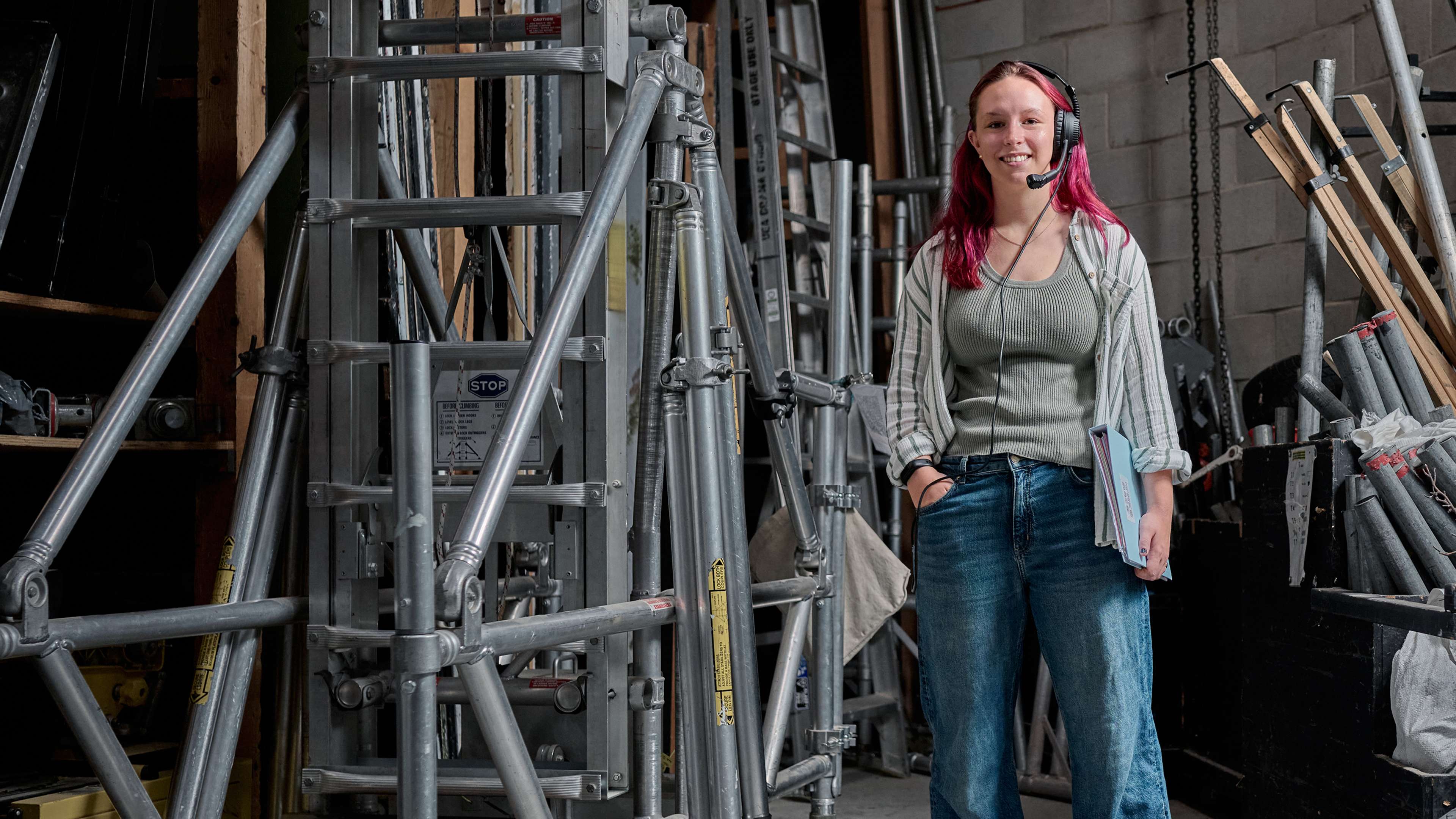 A student wearing a headset and holding a folder stands backstage and is surrounded by theatre equipment.