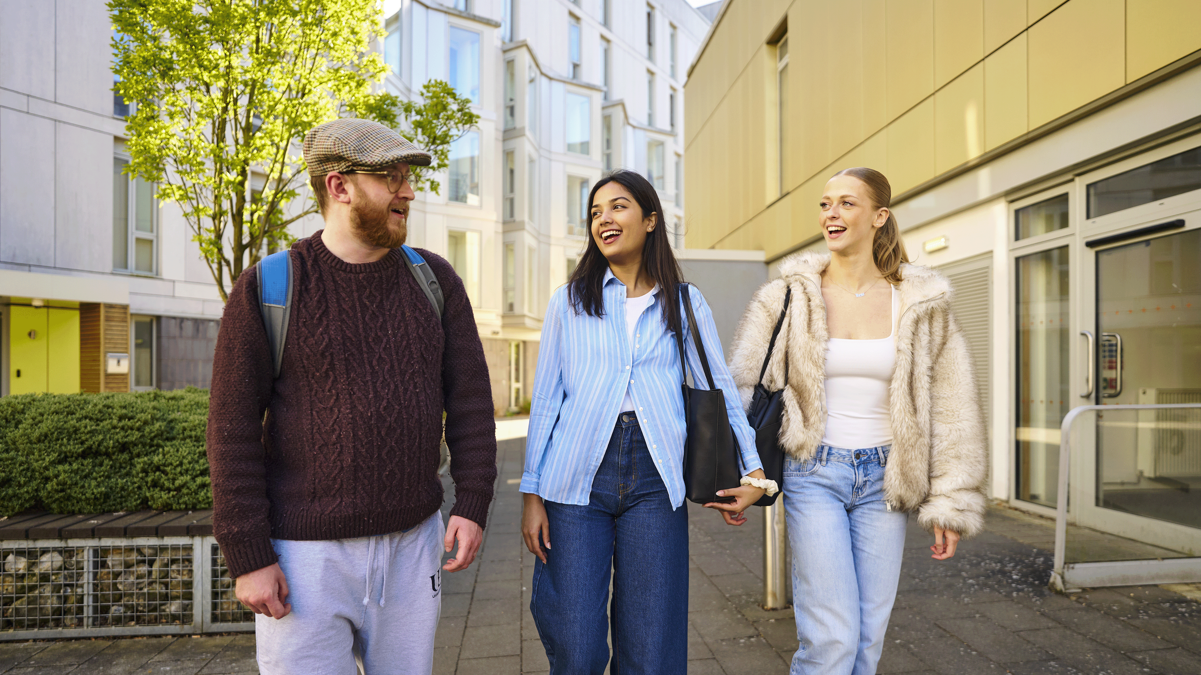 Three smiling students walk along a path outside some campus accommodation.