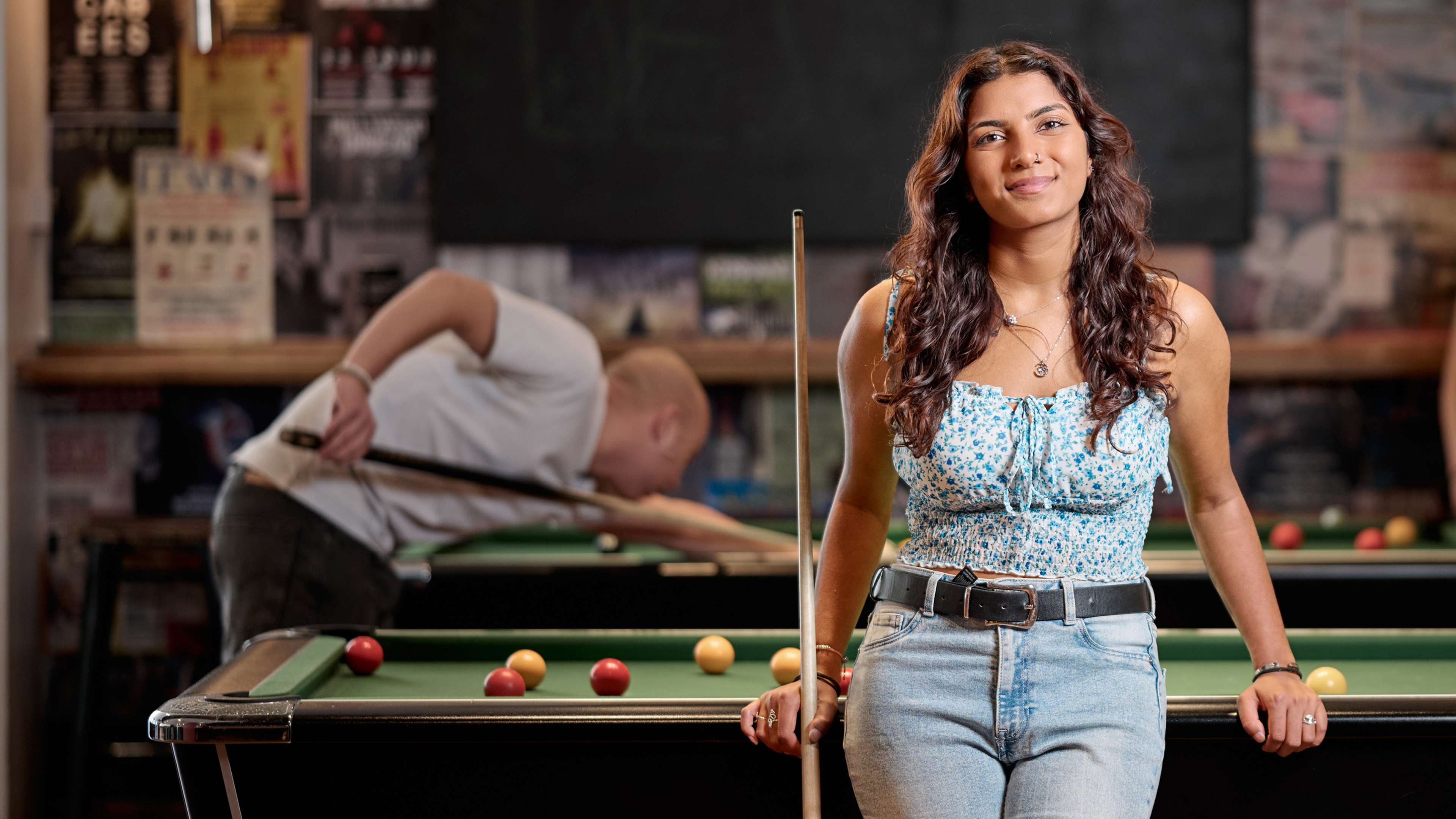 A student holds a pool cue and leans on a pool table. She is smiling at the camera.