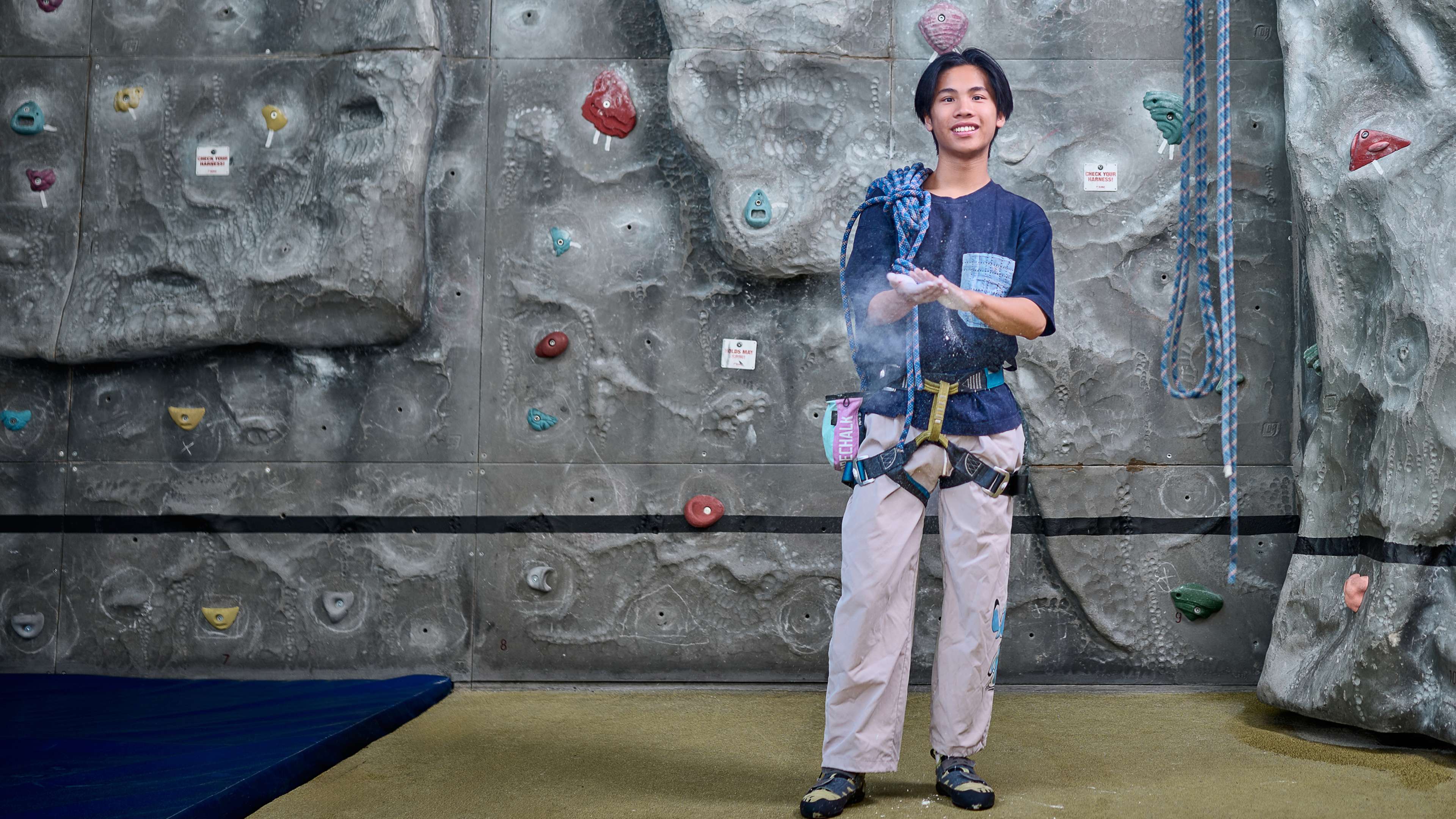 A student wearing climbing equipment stands in front of a climbing wall. He is putting chalk on his hands.