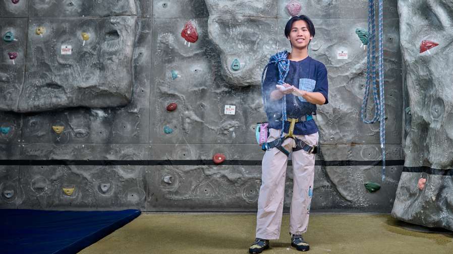 A student wearing climbing equipment stands in front of a climbing wall. He is putting chalk on his hands.
