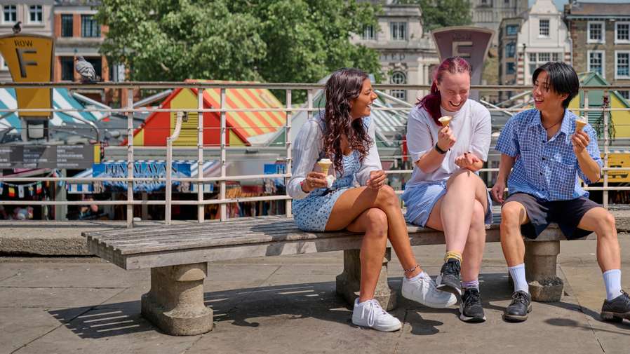 Three students sit on a bench eating ice cream near Norwich market. Norwich castle can be seen in background.