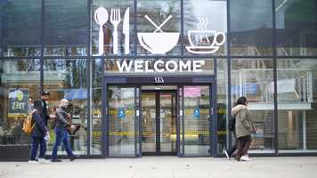 A group of students walking in front of a building with full length glass windows, the automatic doors are open and above the doors it says 'WELCOME' and above that are, left to right, pictures of cutlery, a bowl with noodles sticking out the top, and a steaming cup and saucer with UEA inside the cup silhouette