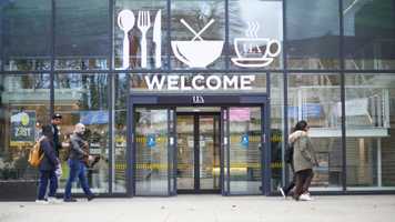 A group of students walking in front of a building with full length glass windows, the automatic doors are open and above the doors it says 'WELCOME' and above that are, left to right, pictures of cutlery, a bowl with noodles sticking out the top, and a steaming cup and saucer with UEA inside the cup silhouette