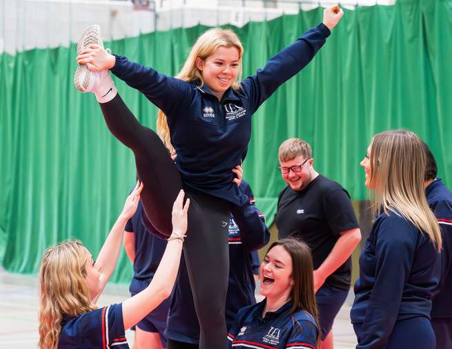 Students practice a cheerleading stunt indoors, with one lifted into the air on one leg, arms raised in a 'V', while teammates observe and support.