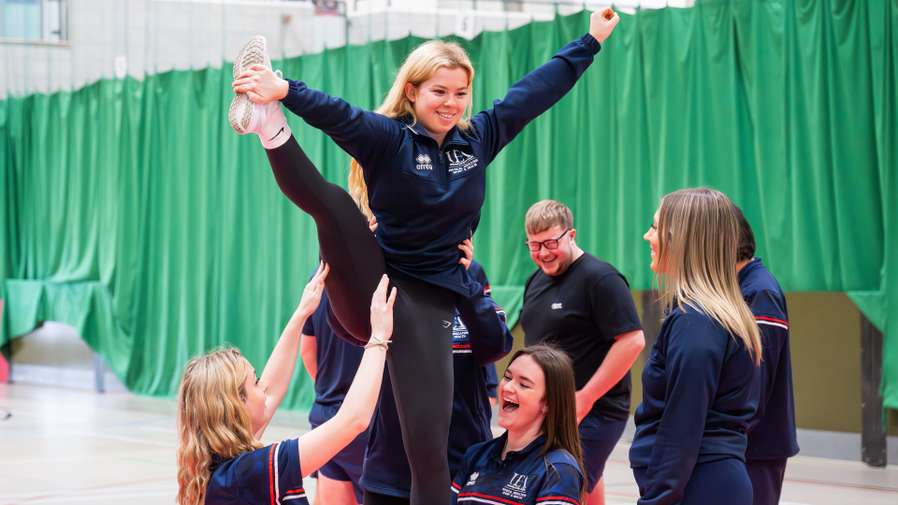 Students practice a cheerleading stunt indoors, with one lifted into the air on one leg, arms raised in a 'V', while teammates observe and support.