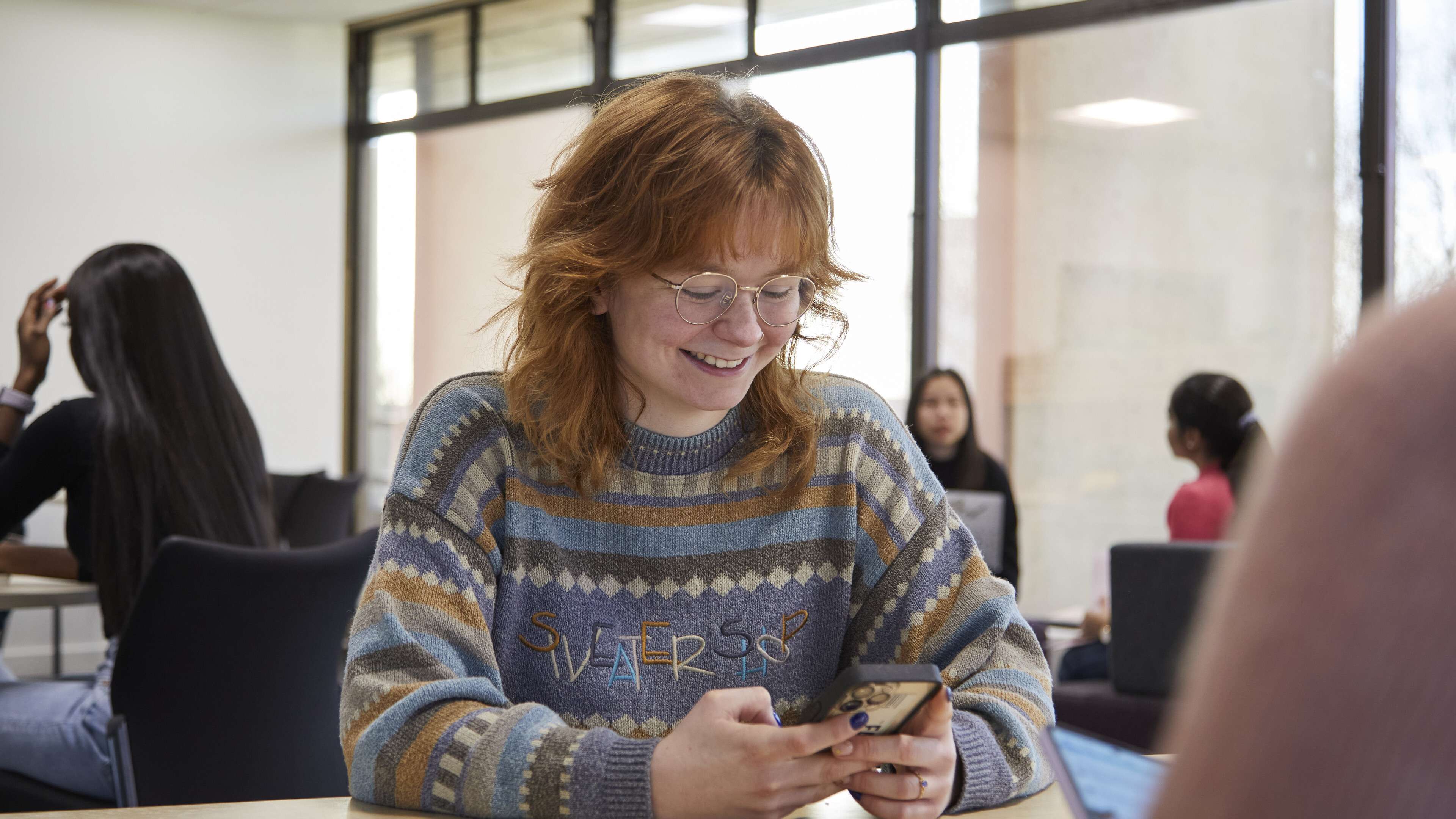 A smiling student with red hair and glasses uses her phone at a desk in a bright university classroom, with other students in the background.