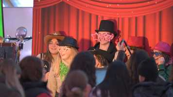 A group of students standing in front of a red background in fancy dress props to take a photobooth picture.