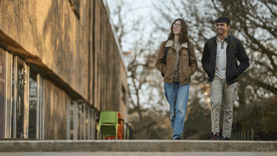 Two smiling students walk along a path outside a building.