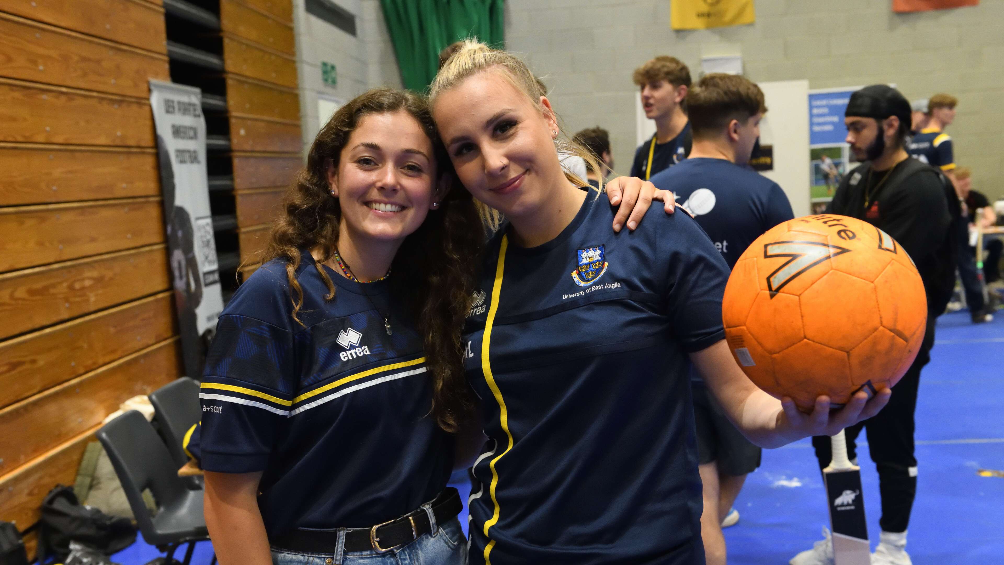 Two students in a sports hall smiling into the camera, one holding a volleyball.