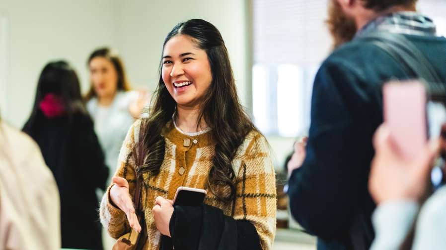 A student with long dark hair and wearing a yellow cardigan is smiling in a group setting. They are looking just past the camera in a candid moment and looks happy and confident. Other people are visible in the background.