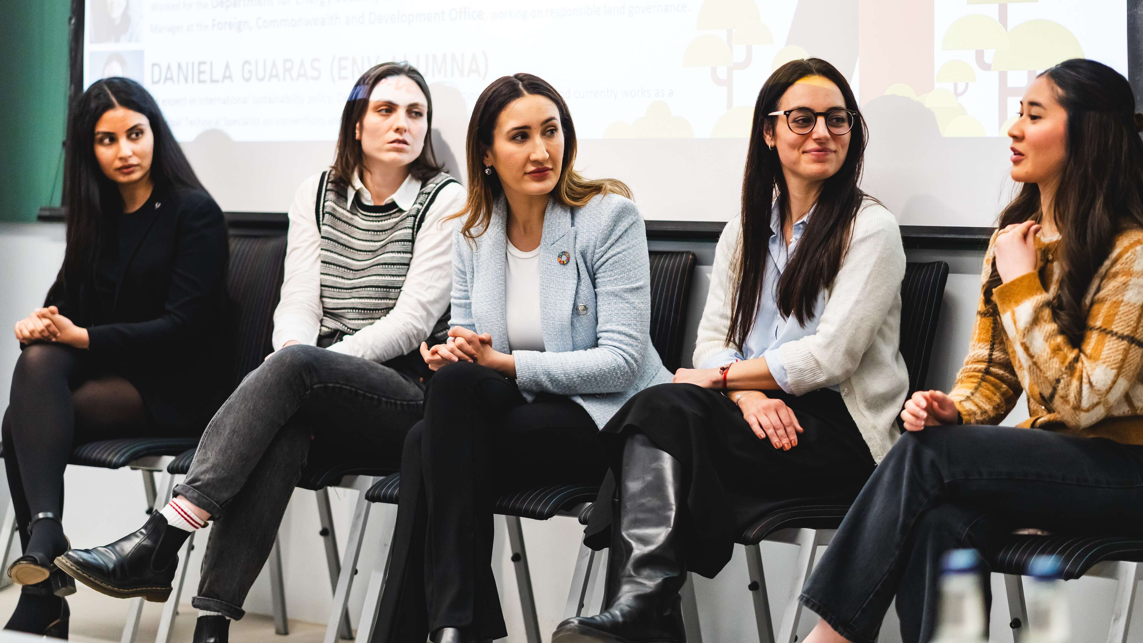 Five women sitting on chairs, engaged in a panel discussion in a conference room. A presentation screen is visible in the background.