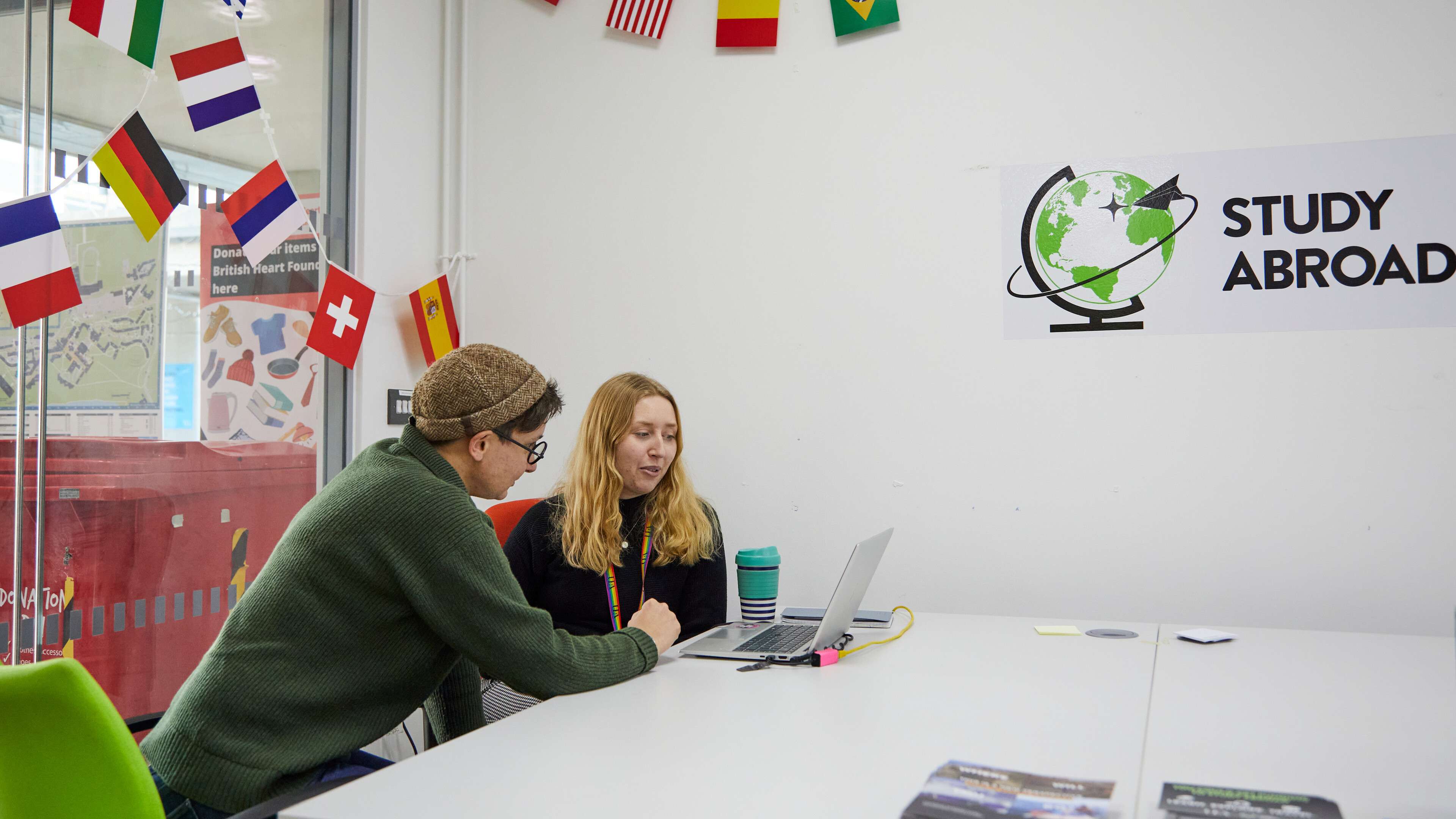 Two people looking at a laptop in front of various international flags.