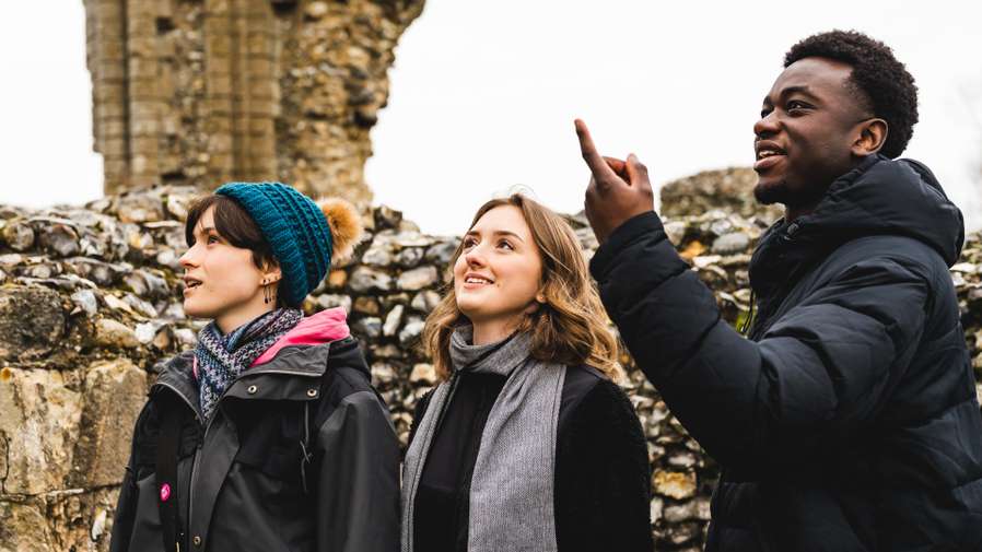 Three people in winter clothing admire historic stone ruins, with one pointing upwards.