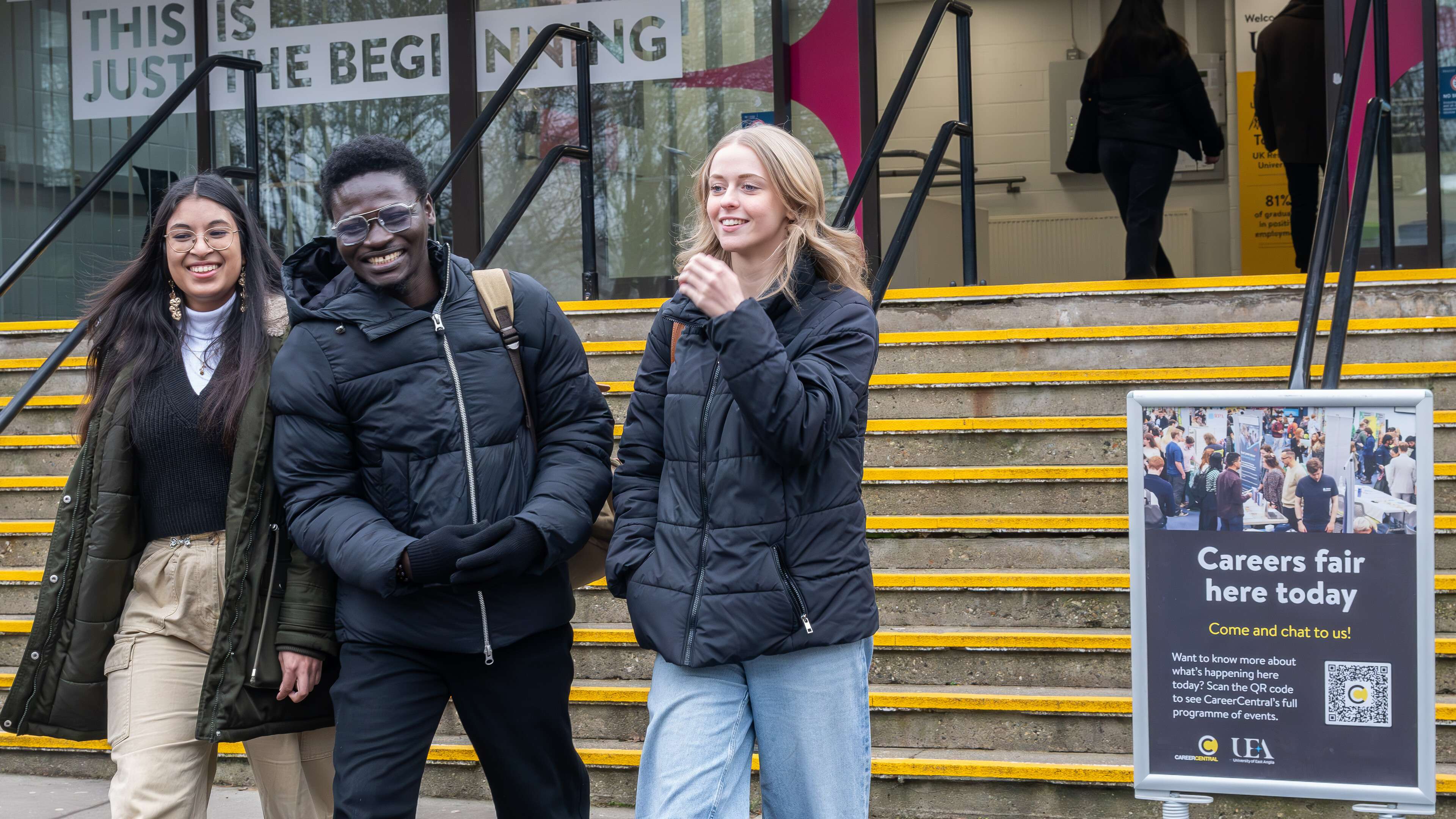 Three smiling students in winter coats walk down steps outside the UEA Congregation Hall, with a sign advertising a careers fair nearby. The sign encourages students to attend and includes a QR code for event details. A banner above the entrance reads 'This is just the beginning.'