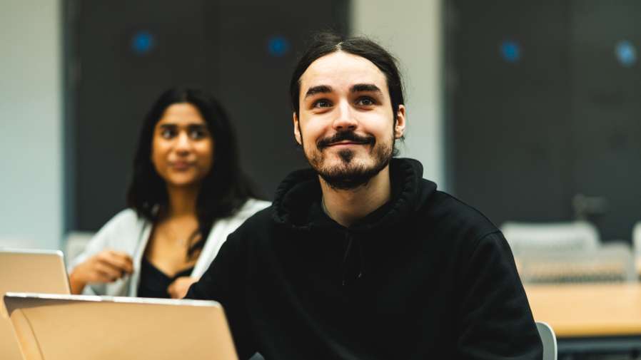Two people in a classroom setting, one smiling in the foreground with a laptop, and another slightly blurred in the background.