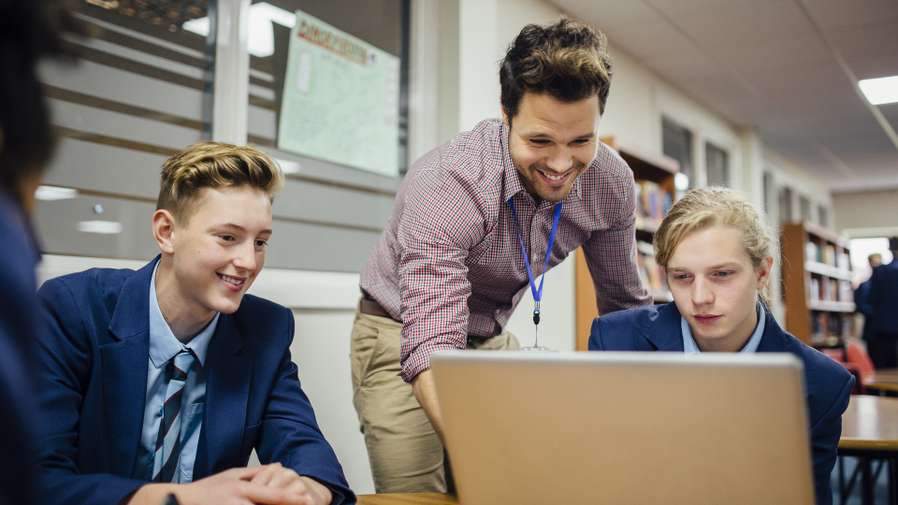 Teen students are working on a laptop in their school lesson. A teacher is leaning over them, helping them with some work. Keywords: