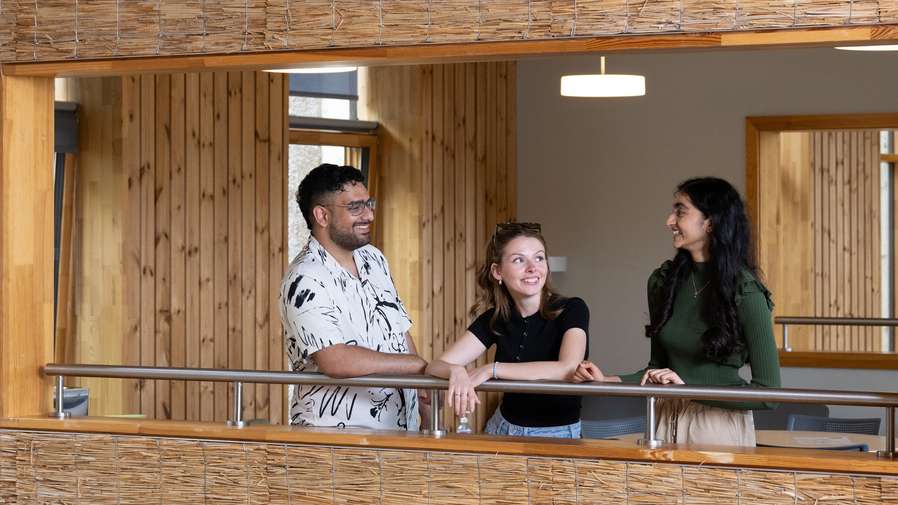 Three studetns on a balcony in the UEA Enterprise centre. They are framed in a window, leaning on the railing of a balcony and chatting. The stuent on the left is wearing a white graphic-patterned shirt, the student n the middle has sunglasses on her head and a black t-shirt, and the student on the right is in profile, wearing a high-necked green top with long sleeves.