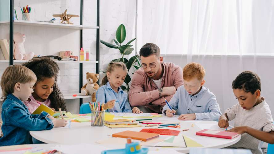 teacher and interracial preschoolers at table with paints and papers in classroom