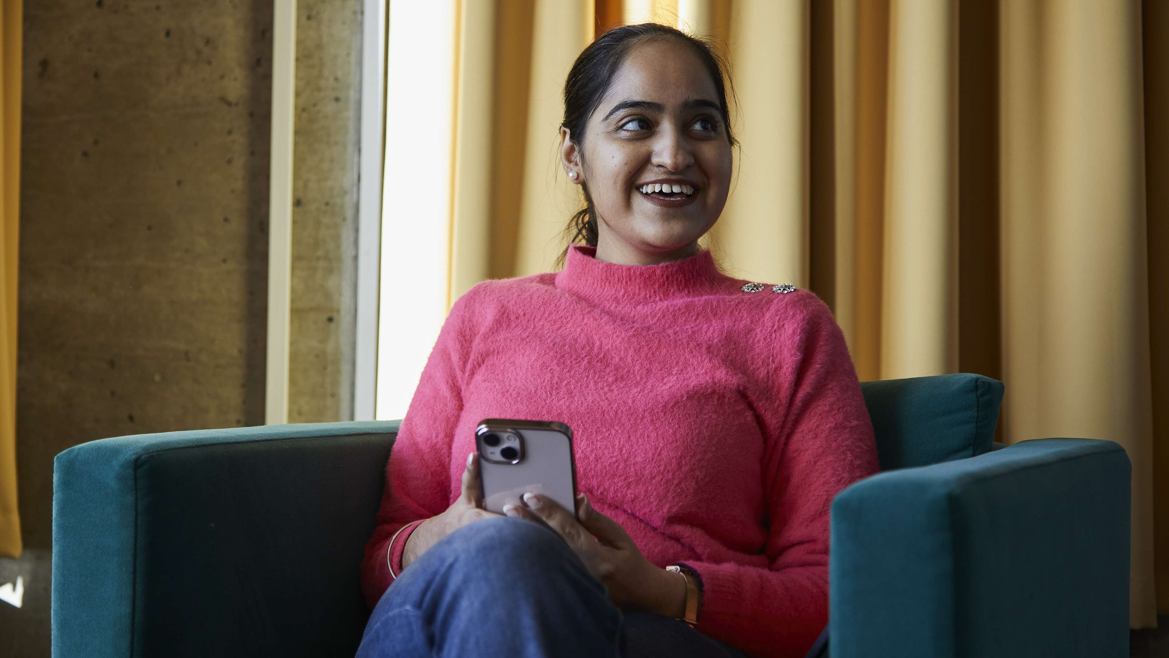 A female student smiles while sitting in a modern university common room, wearing a pink jumper and holding her phone as she looks to her right.