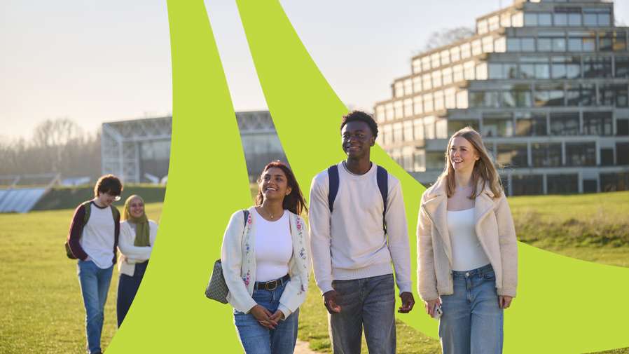 A diverse group of students smiling while walking on a sunny university campus in Norwich, with modern buildings in the background.