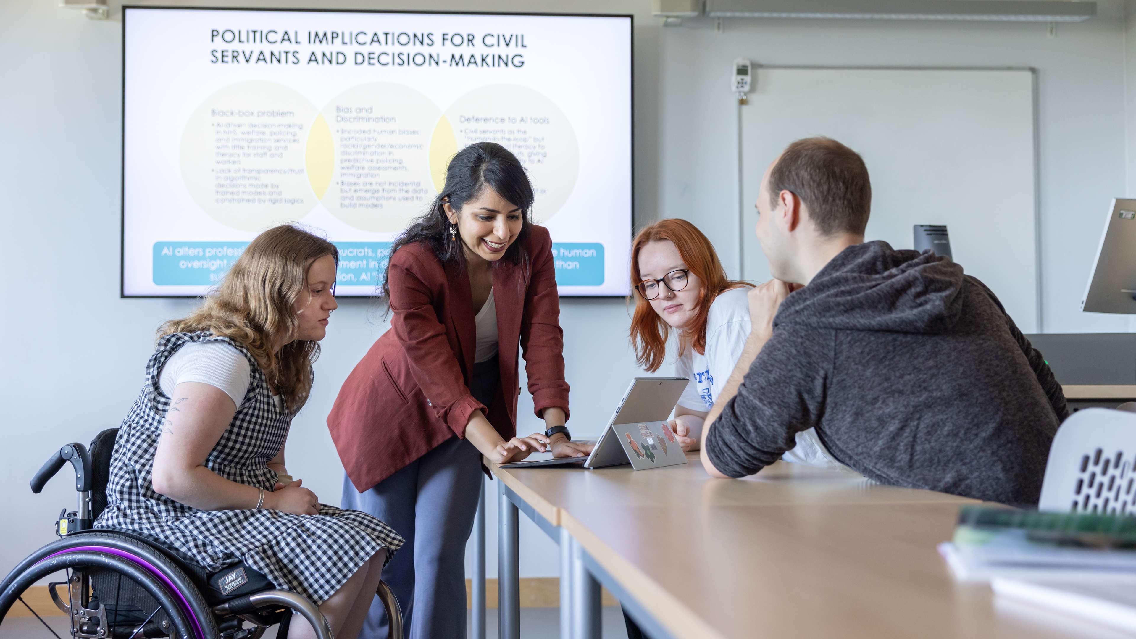 Three students and a lecturer looking at a laptop on the table - the lecturer is stood up and one student is in a wheelchair. There is teaching material on the screen behind them
