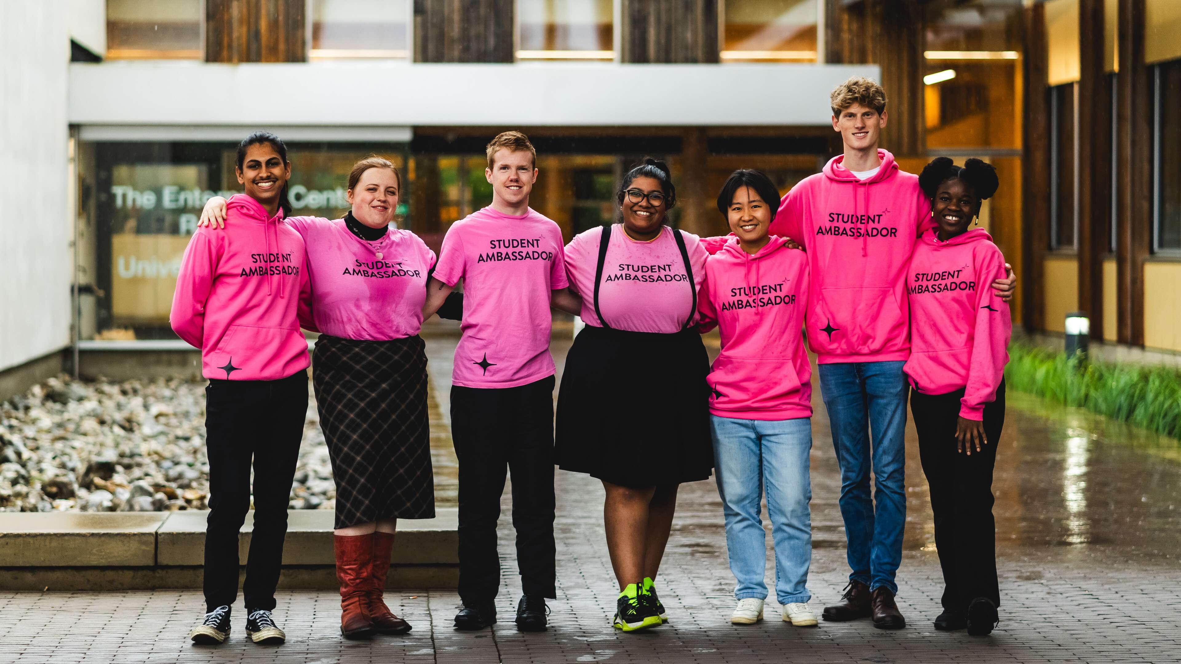 A group of seven UEA student ambassadors wearing matching bright pink hoodies and t-shirts stand together outside the UEA Enterprise Centre, smiling with their arms around each other