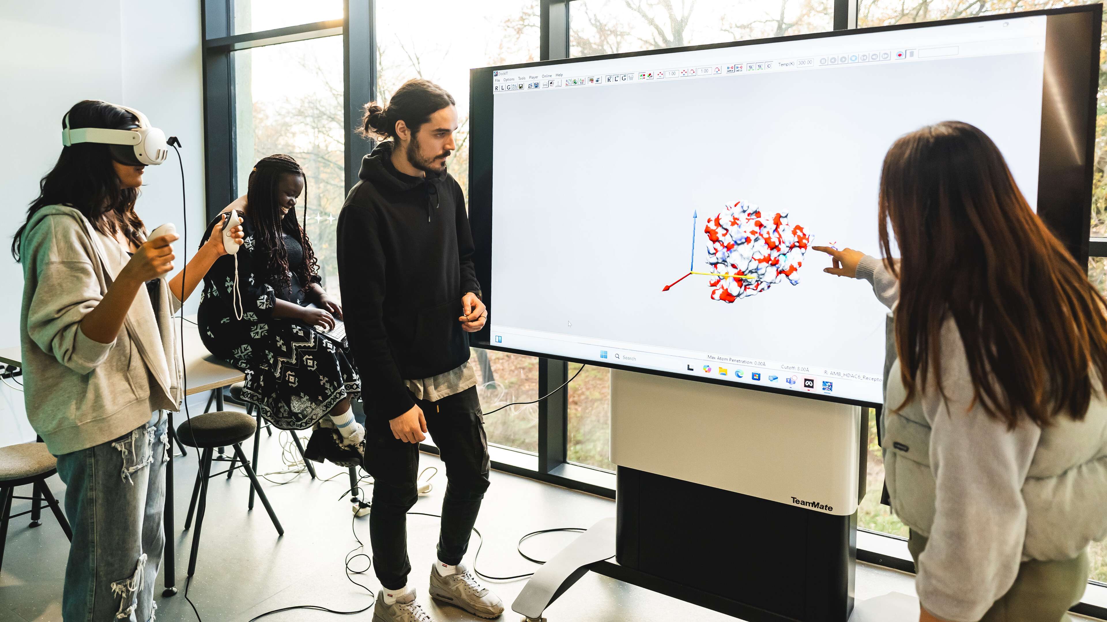 Four people in a room, three engaging with a large interactive screen displaying a colorful 3D model. One wears a VR headset.
