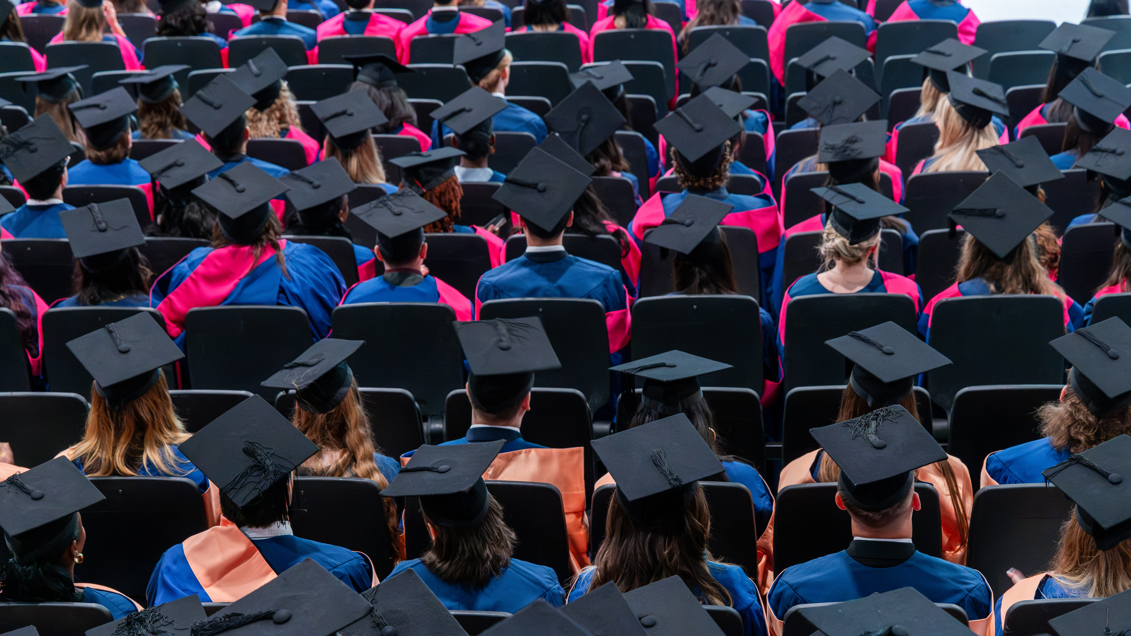 A sea of graduates in grad caps and gowns fills an auditorium during a university graduation ceremony in Norwich.