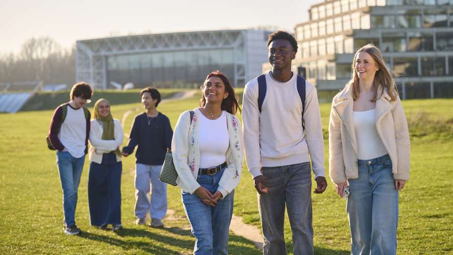 Six students walk across the UEA campus on a sunny day. There are three students in the foreground and three in the back. They are walking into the front of the shot and looking around, chatting. The Sainsbury Centre is behind them