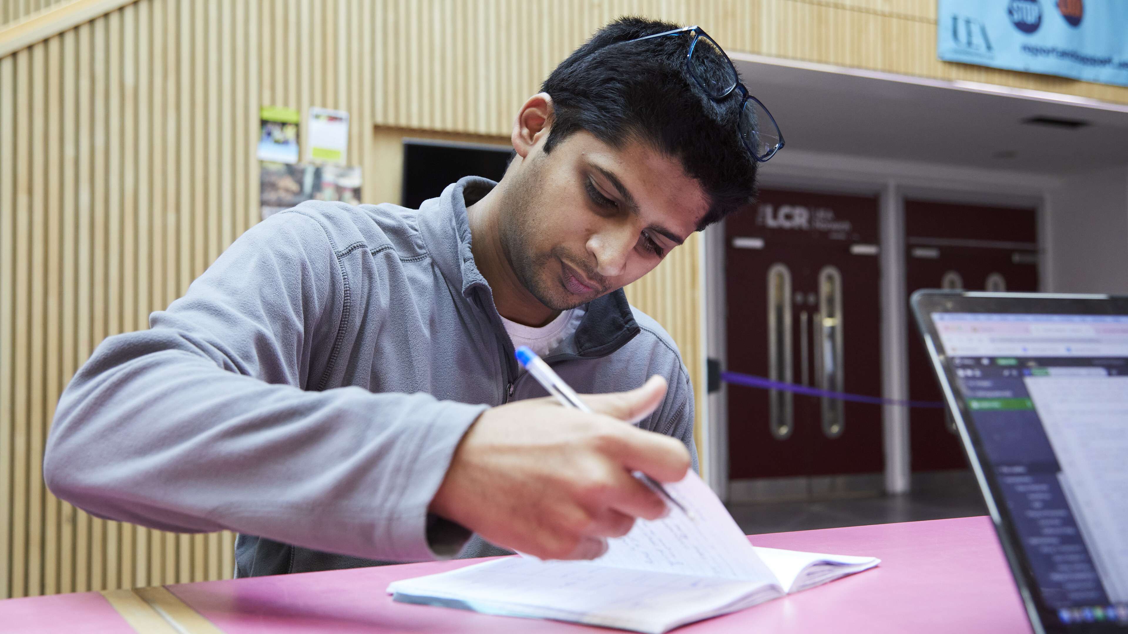 A student sits at a desk, writing notes in a notebook with his laptop open nearby.