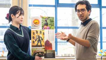 Two students from the school of Global Development present a colorful poster with various images and text in a bright room with large windows.