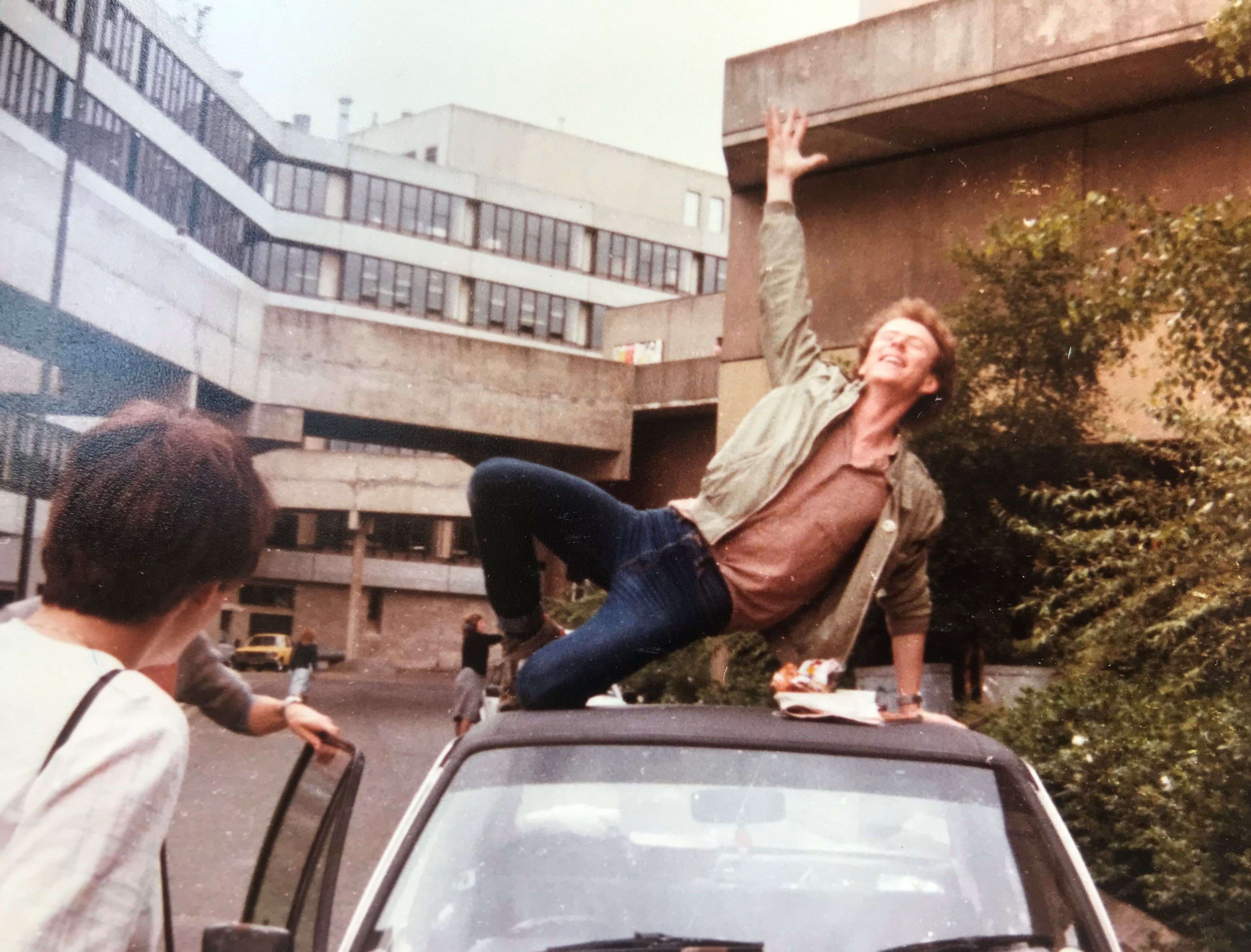 A picture of Andrew on top of his MkII Ford Escort outside his room in Norfolk Terrace. His right arm is raised above him and he carries a broad smile.