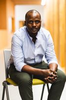Man sat leaning forward in chair in blue shirt and khaki trousers with hands clasped