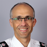 Man with glasses smiling, wearing a white uniform with black and red insignia on the shoulders, against a grey background.