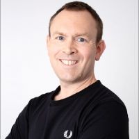 Smiling person with short hair wearing a black shirt, standing against a plain white background.