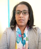 A person wearing glasses, a floral scarf, and a gray blazer, standing against a light-colored background.
