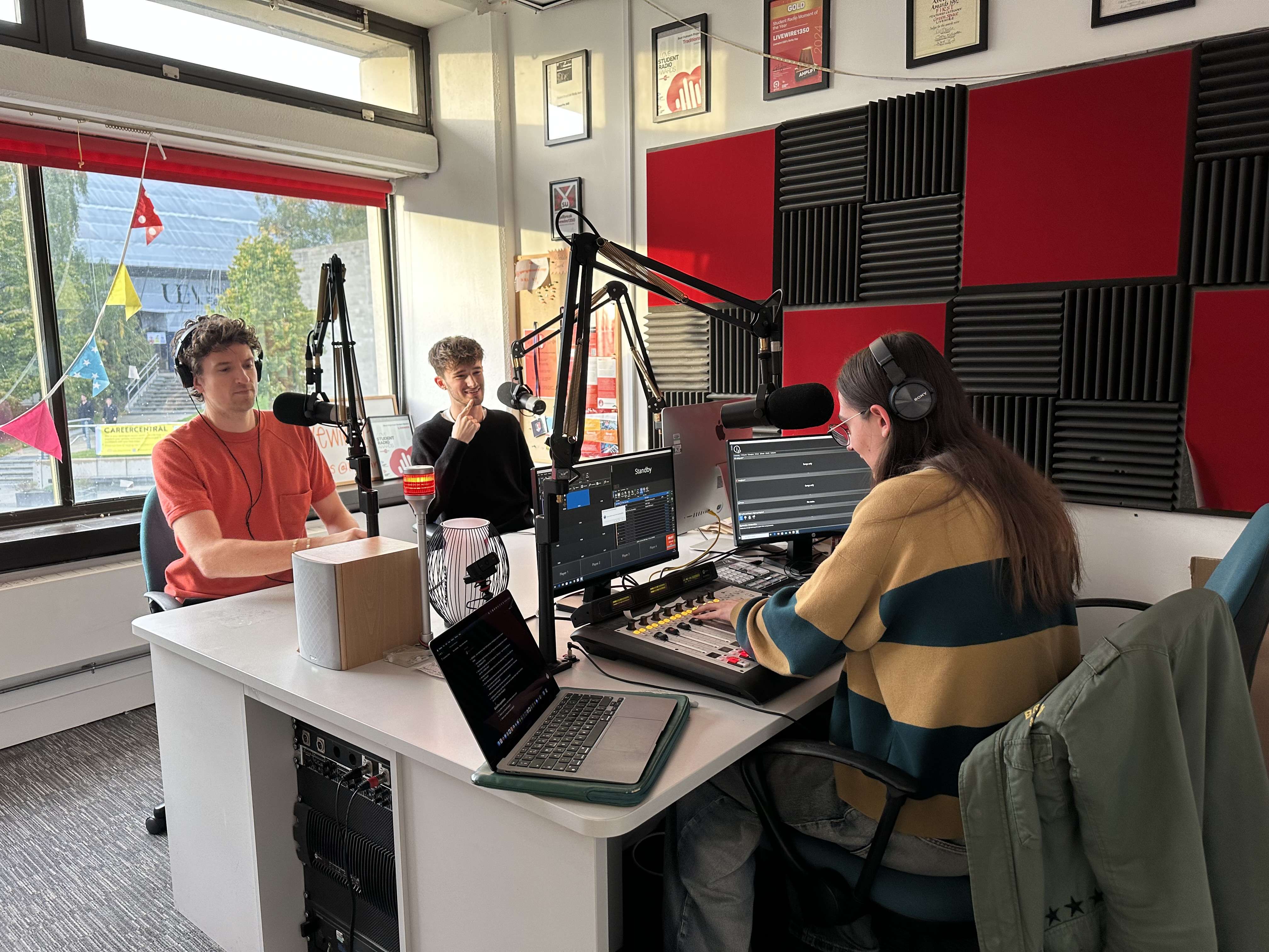 Three people seated in a room with microphones and recording equipment. A red and black checkerboard wall is to their right.