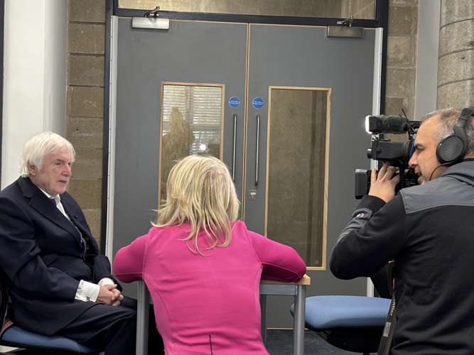 Old man sat at table facing woman who has her back to the camera next to cameraman facing old man