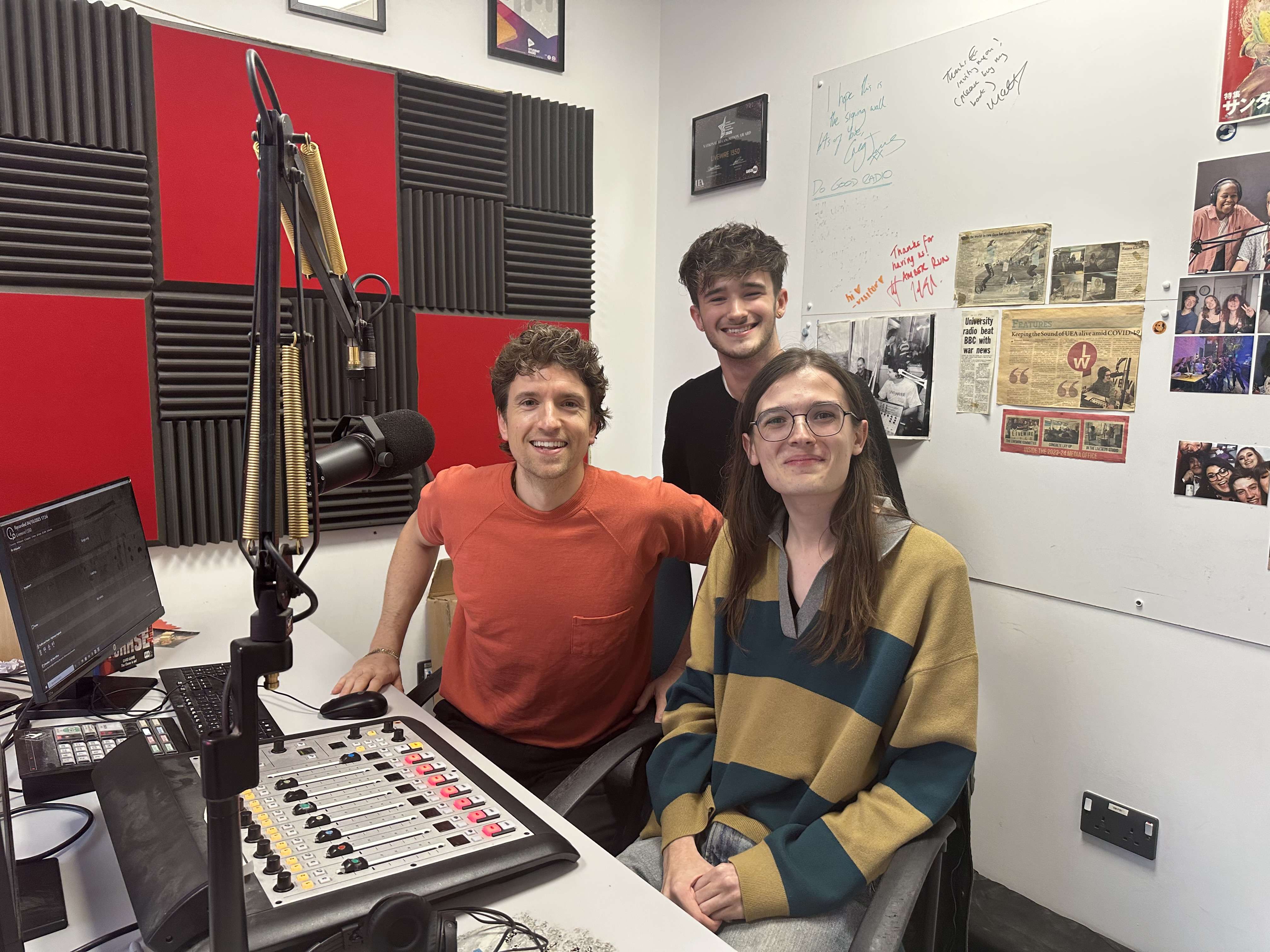 Three people smiling at the camera in a radio recording room. A wall of newspaper clippings is in the background and a red and black checkerboard wall to the left.