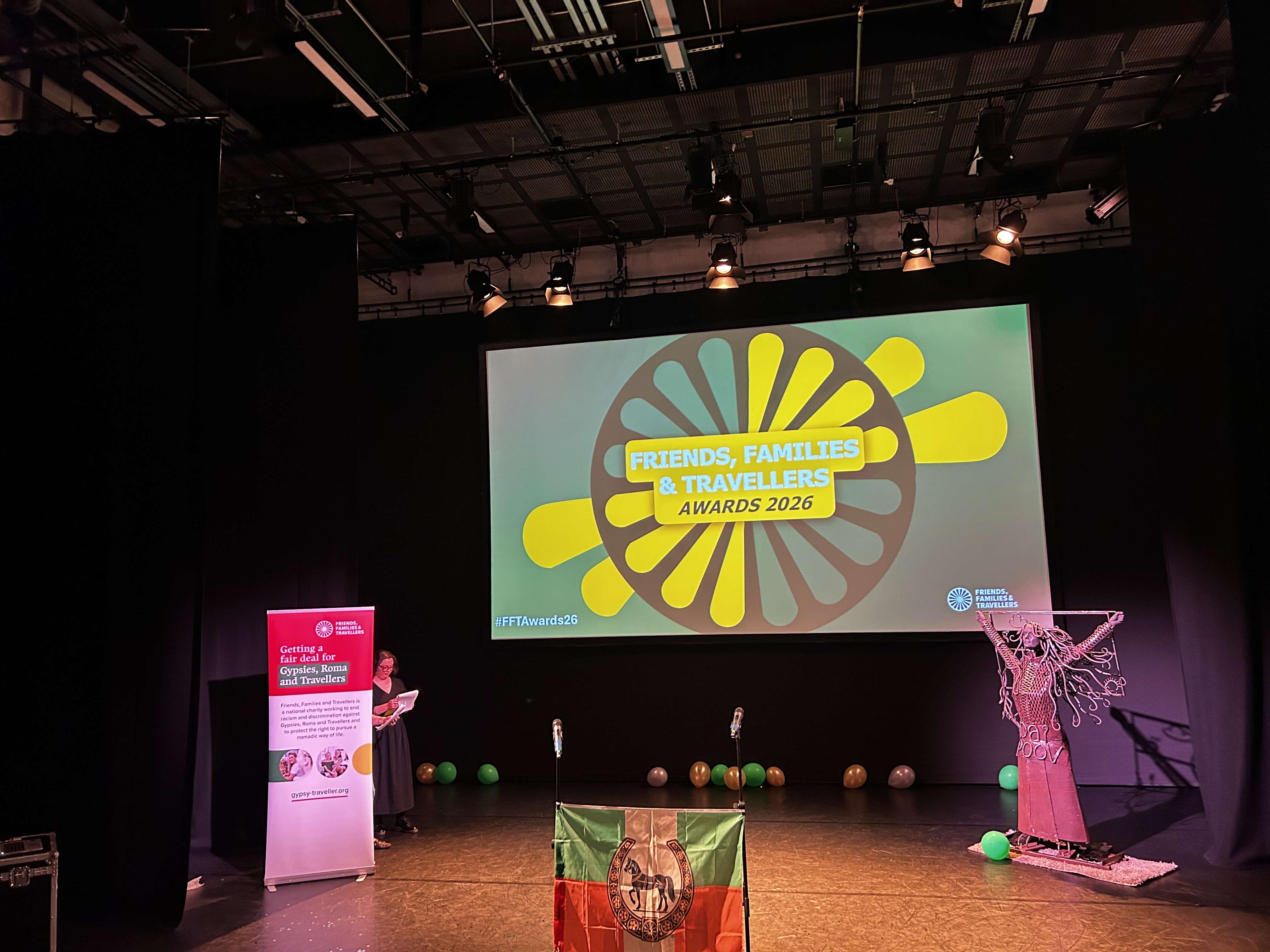 Event logo displayed on stage for the Friends, Families & Travellers Awards with a Romani flag in the foreground.